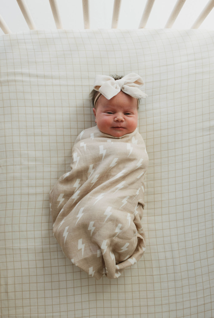 A newborn baby is swaddled in a Forever French Baby Muslin Swaddle with tan and cream lightning bolt patterns, lying on a beige and white checkered sheet. The baby is wearing a beige headband with a large bow and has a content expression. The background shows the slats of a crib.