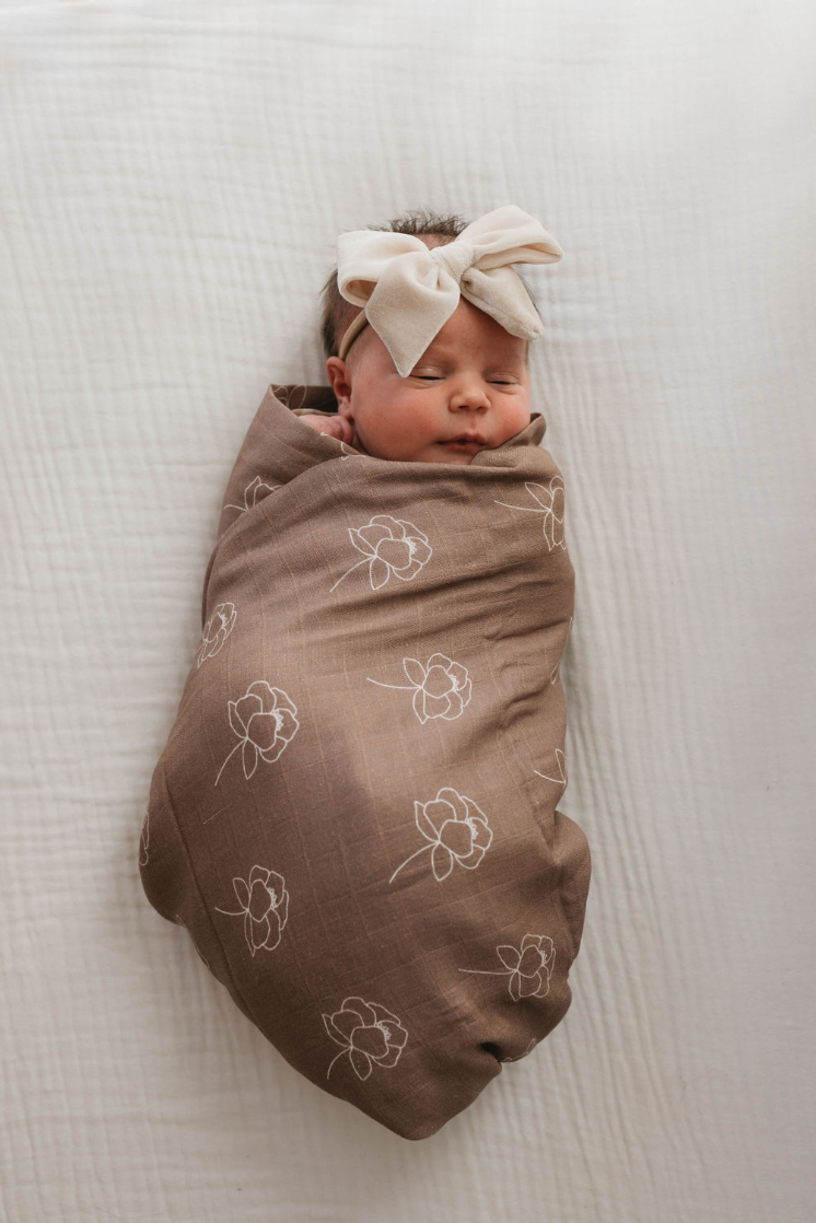 A newborn baby is wrapped in a forever french baby's Muslin Swaddle | Sweet Dreams Floral, lying on a white surface. The baby is asleep and has a large beige bow headband on their head.