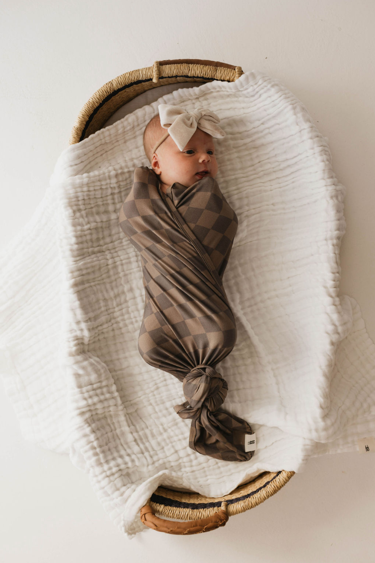 A baby, swaddled in a "Faded Brown Checkerboard" Bamboo Swaddle from Forever French Baby and wearing a beige headband with a bow, lies in a wicker basket on a soft white blanket. The baby is gazing to the side, creating a peaceful and cozy scene.