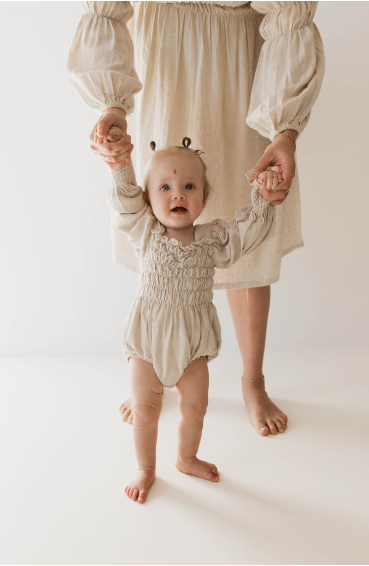 A baby dressed in a luxurious Linen Romper in Flax by forever french baby is standing and holding hands with an adult, who is partially visible and wearing a matching beige dress. The baby, dressed in the Flax Long Sleeve Linen Romper available in sizes 0-24 months, has a small red mark on their forehead and is looking at the camera.