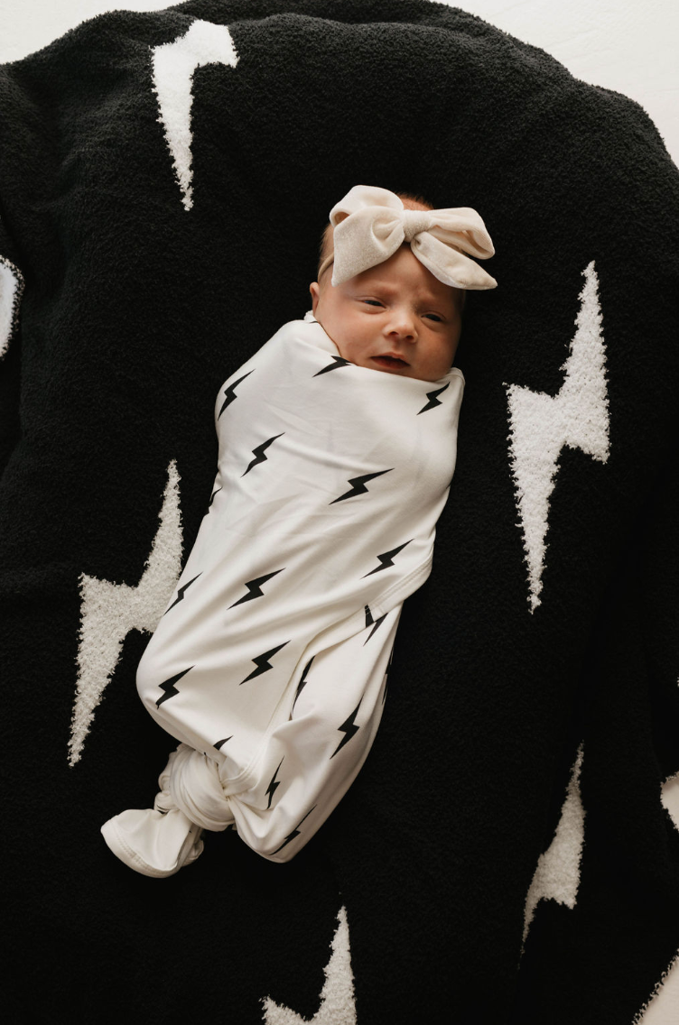 A baby wrapped in the forever french baby Bamboo Swaddle | White & Black Lightning Bolt lies on a black blanket with matching white lightning bolt patterns. The baby wears a light-colored bow headband and appears calm and relaxed, thanks to the breathable sleepwear keeping them comfortable.