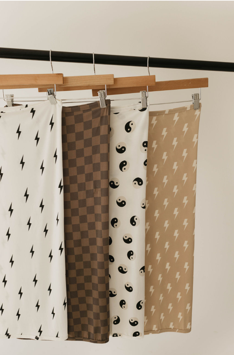 Four patterned fabrics hang on a wooden rack. From left to right: white fabric with black lightning bolts, faded brown checkerboard bamboo swaddle from forever french baby, white fabric with black yin-yang symbols, and beige fabric with white lightning bolts. Ideal for bamboo swaddles or baby sleepwear due to their hypo-allergenic qualities.