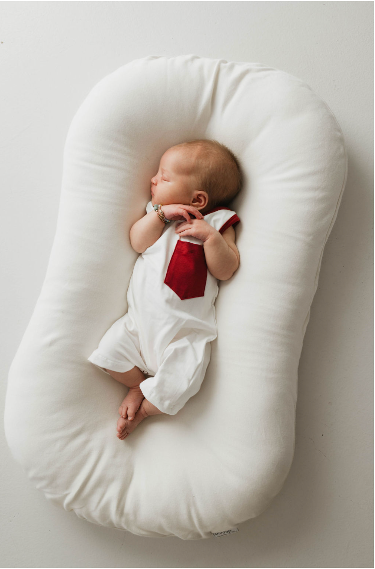 A newborn baby lying on a white pillow wears the "T-Shirt Tank Romper w/ Pocket | White & Red" by forever french baby, featuring a red tie design on the front. The baby's hands are close to its face, with a small bracelet on one wrist. Perfect for warmer weather, this outfit simplifies diaper changes against the plain white background.