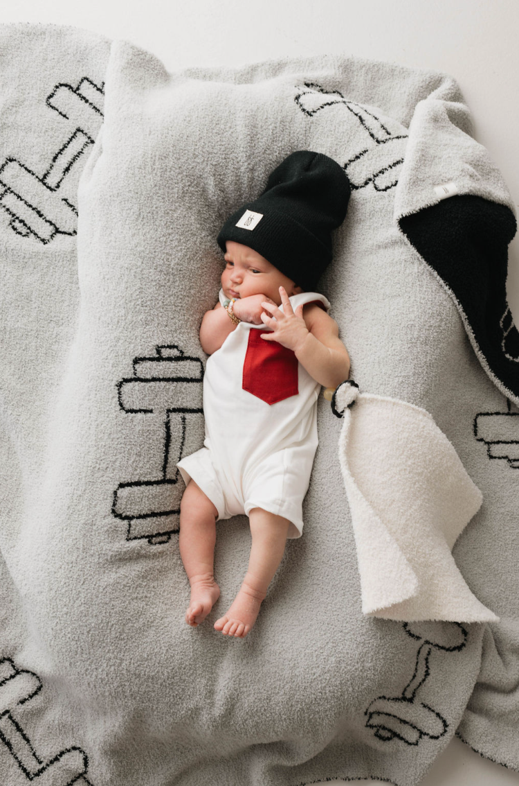 A baby dressed in a "T-Shirt Tank Romper w/ Pocket | White & Red" from forever french baby and a black beanie lies on a soft, light grey blanket adorned with black and white birds. Perfect for warmer weather, the lightweight romper allows easy diaper changes. The baby is raising both hands near their mouth.