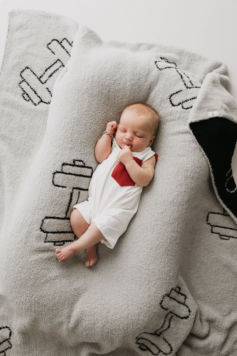 A sleeping baby dressed in a forever french baby's T-Shirt Tank Romper w/ Pocket | White & Red rests on a light gray blanket decorated with black-and-white barbell patterns. The baby is lying on its back, with one hand near its face, positioned peacefully.