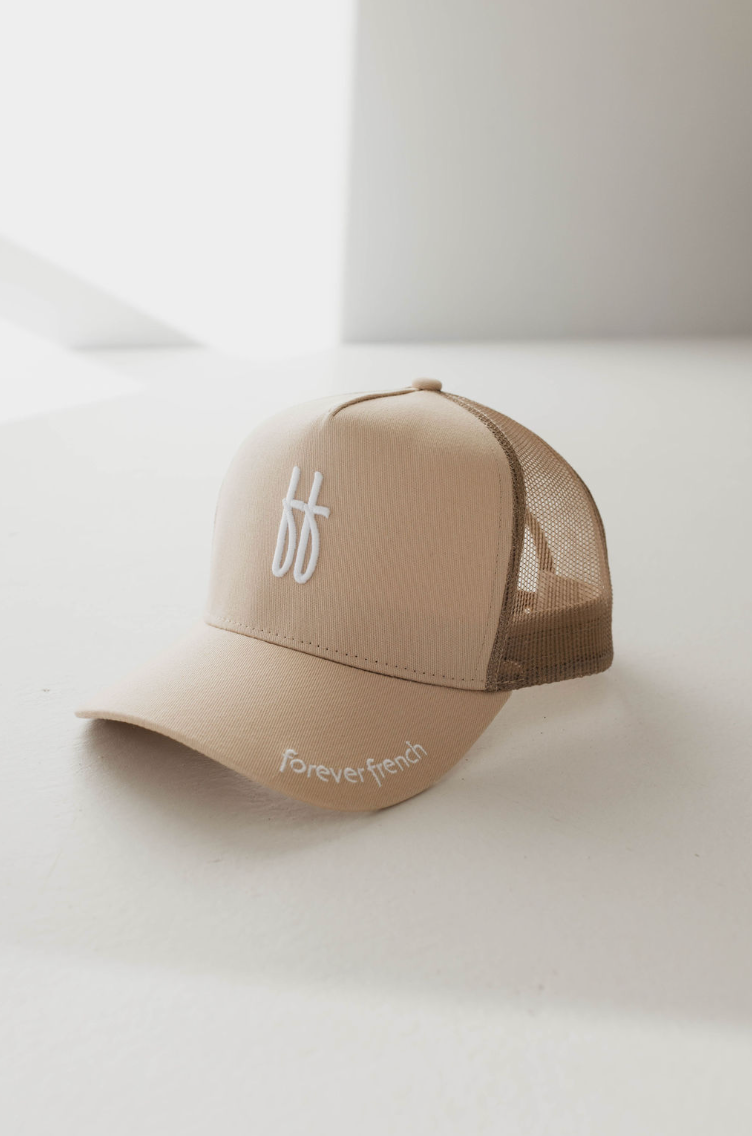 A tan trucker cap with a brown mesh back and a curved brim. The front features a white "ff" logo, while the brim displays "Forever French" embroidered in white on the left side. Ideal for spot cleaning with warm water, this Adult Trucker Hat from forever french baby (model: ff Blackout) is resting on a white surface under soft lighting.