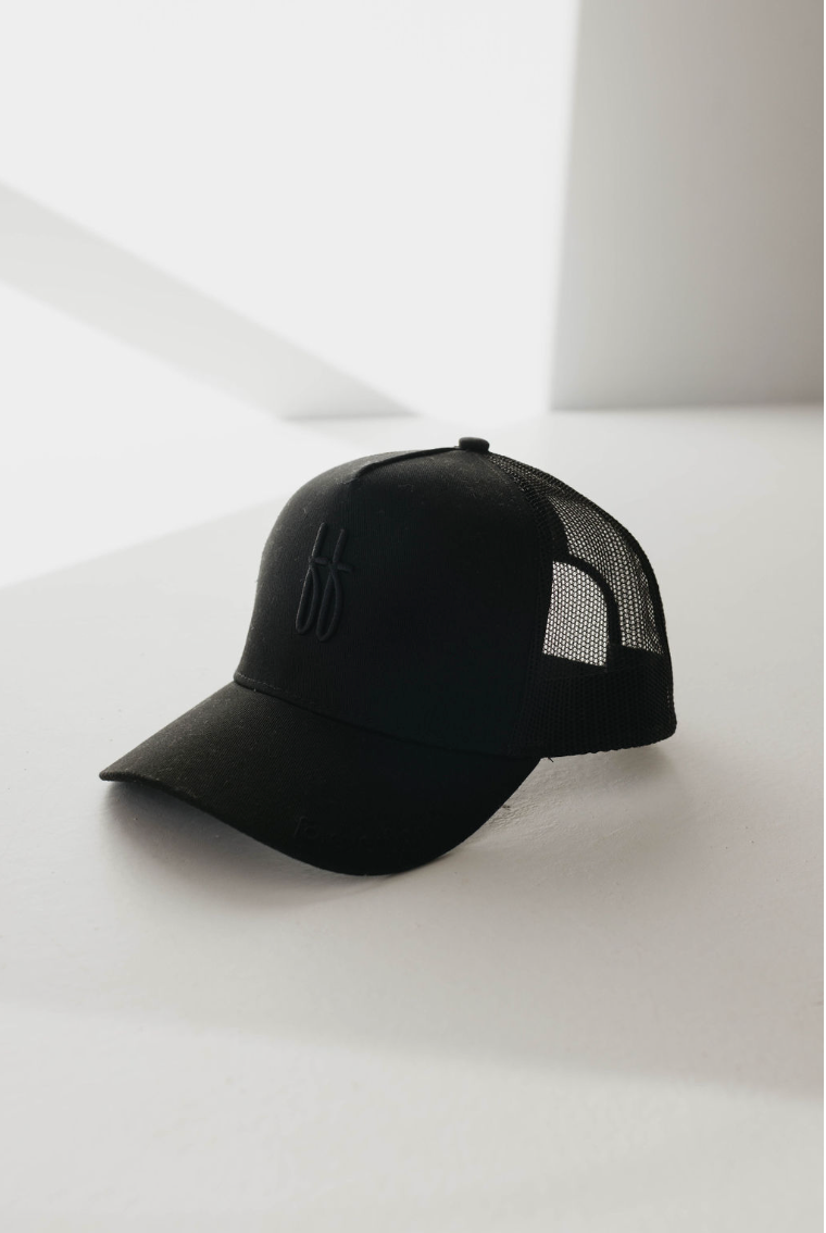 The Adult Trucker Hat | ff Blackout by forever french baby is displayed on a white surface with sunlight and shadow in the background. The front of the black cap features a subtle, embroidered design, while its mesh back panel showcases the classic trucker hat style. This cap is ideal for spot cleaning with warm water.