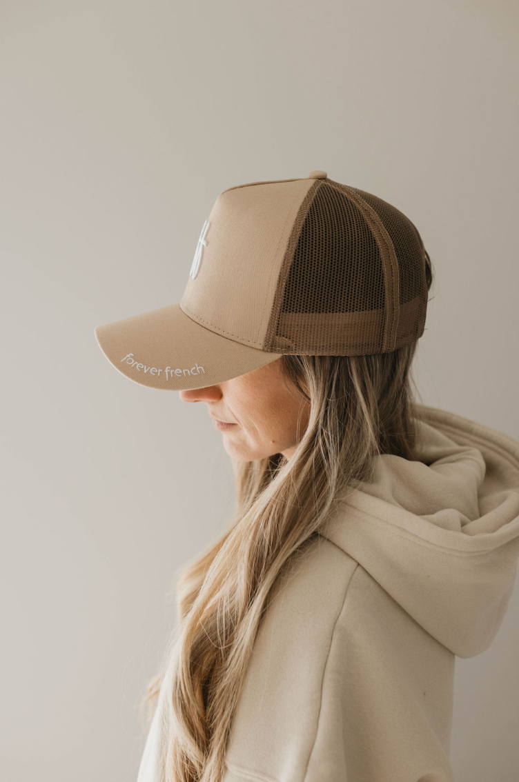 A person with long, blonde hair, wearing the 'Adult Trucker Hat | ff Blackout' by Forever French Baby and a hoodie, is facing sideways against a plain, light-colored backdrop.