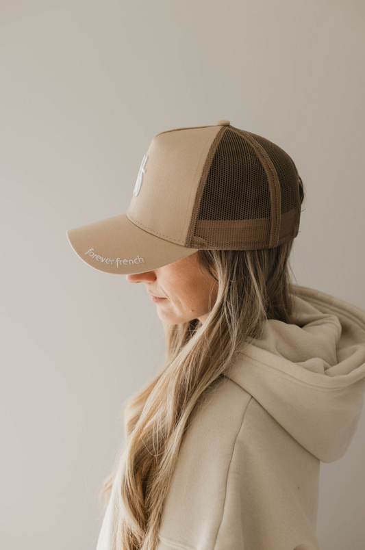 A person with long, blond hair wears a cream-colored hoodie and an Adult Trucker Hat | ff Tan by forever french baby. They face sideways, showcasing the cap's profile and their relaxed posture against a neutral background.