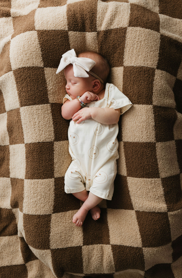 A sleeping baby in a white outfit adorned with yellow smiley faces and a large white bow headband lies on the Brown Sugar Checkerboard plush blanket by forever french baby. The baby is positioned diagonally, with arms and legs slightly curled, enveloped in the cozy comfort of microfiber feather yarn adhering to OEKO Standard 100.