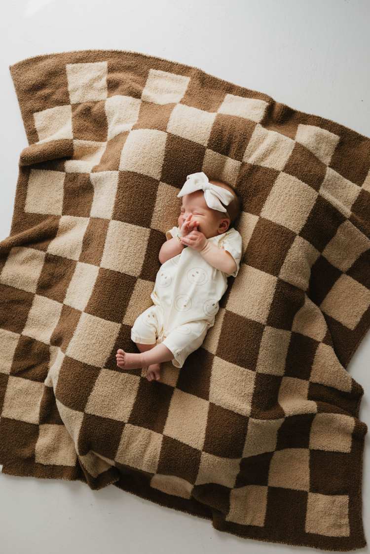 A baby, dressed in a cream outfit and wearing a white headband, lies on the "Brown Sugar Checkerboard" Plush Blanket by forever french baby, made from OEKO Standard 100 microfiber feather yarn. The baby is looking upwards with hands by the face. The soft blanket covers a white surface.