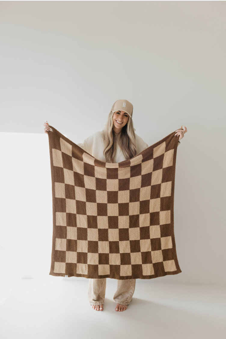 A person with long hair and a cap stands indoors holding the "Plush Blankets | Brown Sugar Checkerboard" by forever french baby. The person is smiling and dressed in light-colored, casual attire against a minimalistic, white background. This luxurious blanket meets the OEKO Standard 100 for safety and quality.