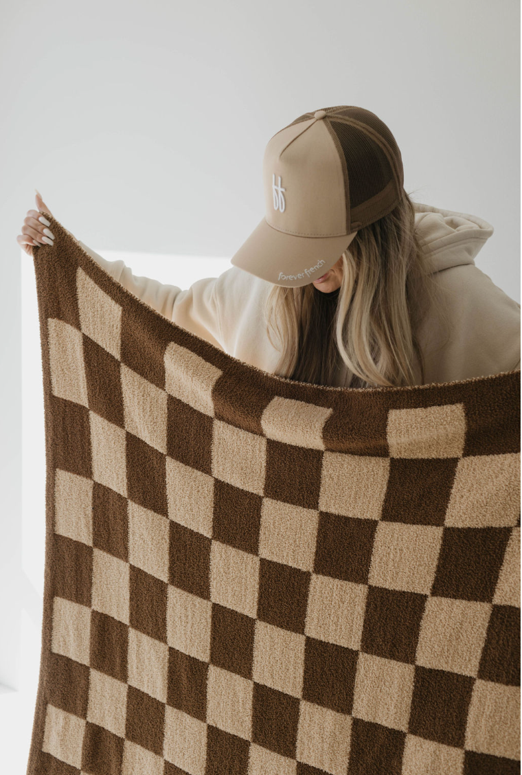 A person wearing a beige and brown trucker hat and a beige hoodie holds up the forever french baby Plush Blankets in Brown Sugar Checkerboard. The background is plain and minimalistic, with soft natural lighting.