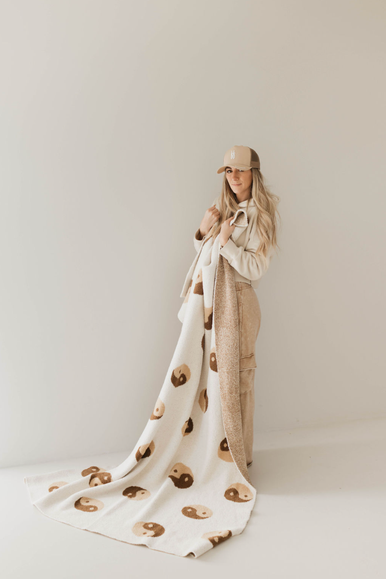 A woman wearing a beige outfit and cap holds a Forever French Baby "Brown Sugar Yin Yang" plush blanket adorned with brown and tan smiley faces. Posing against a simple, light-colored background, the Oeko-tex certified blanket drapes to the floor, adding a cozy and playful touch to the minimalistic setting.
