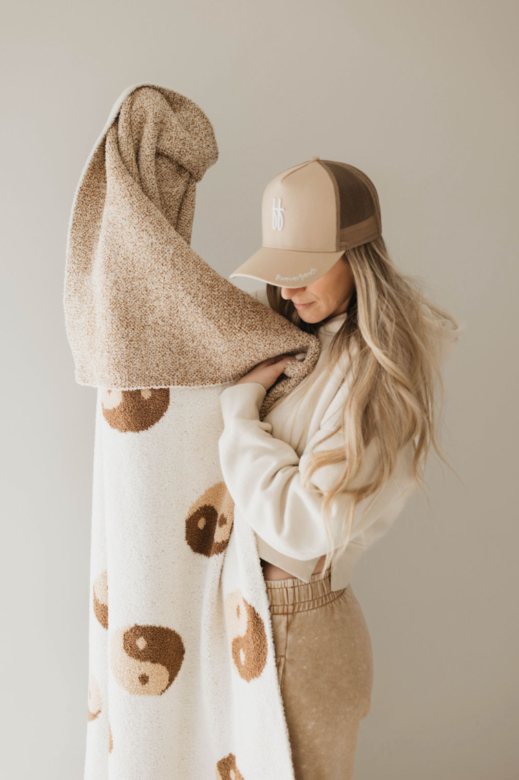 A woman with long blonde hair, wearing a beige and brown baseball cap and neutral-toned clothing, holds an Oeko-tex certified microfiber feather yarn blanket with a bear face pattern. She looks lovingly at the Forever French Baby Brown Sugar Yin Yang Plush Blanket while standing in a minimalistic room with a light-colored background.