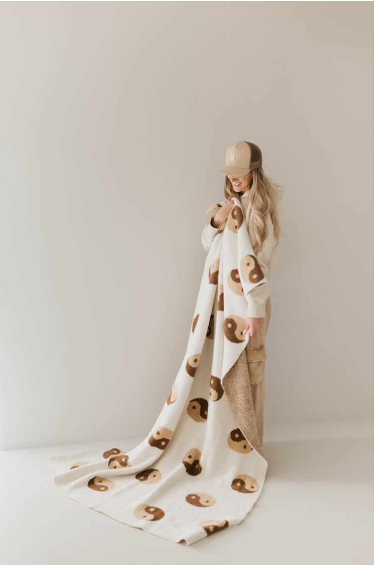 A person with long hair, wearing a beige cap and casual clothes, stands against a plain background. They are holding and partially wrapped in a cozy "Brown Sugar Yin Yang" plush blanket by Forever French Baby, adorned with cute, minimalist bear faces in various shades of brown.