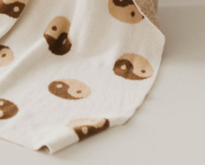 A close-up of the Brown Sugar Yin Yang Plush Blanket by forever french baby, featuring a pattern of smiling face emojis with round, two-tone faces in beige and brown. Some faces are fully visible while others are partially hidden due to the folds in the blanket. Draped over a surface, this delightful piece is reminiscent of the quality found in Oeko-tex certified products.