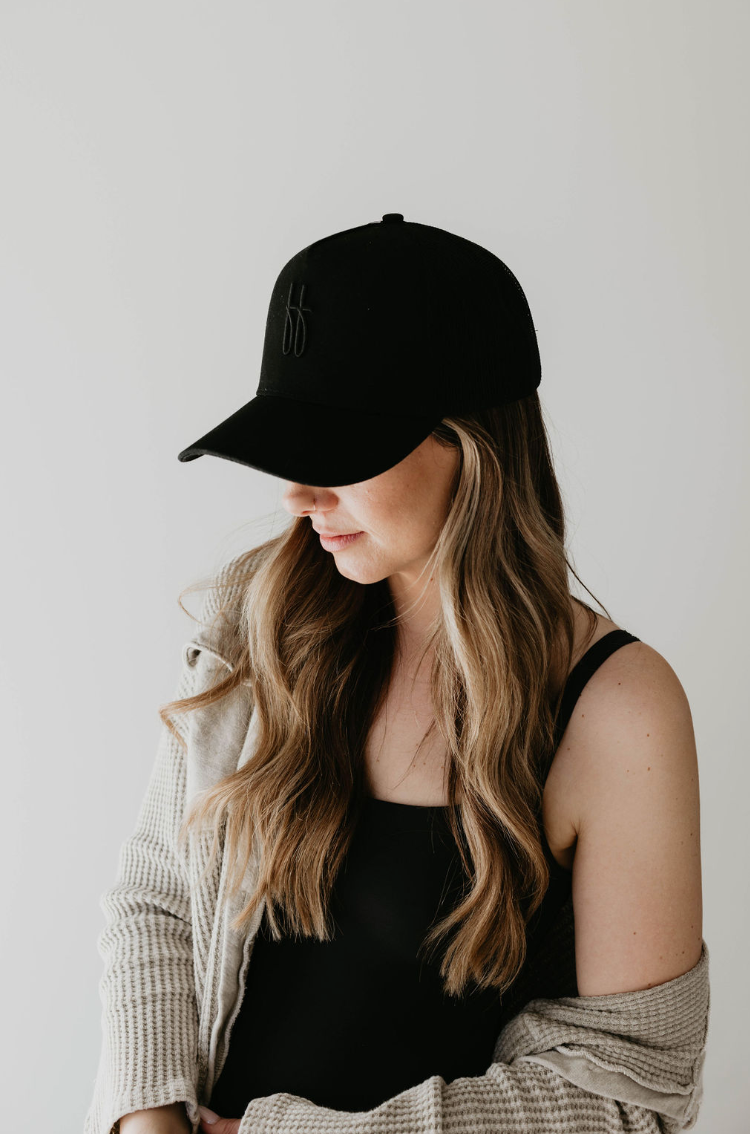 A woman with long, wavy hair is wearing a Forever French Baby Adult Trucker Hat | ff Blackout and a black tank top. A light cardigan drapes over her shoulders as she looks down, partially hiding her face under the cap. The plain, light-colored wall backdrop adds to the effortlessly chic Forever French Baby vibe.