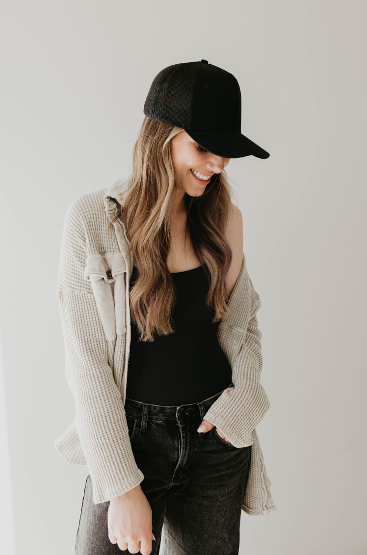 A woman with long, wavy hair is wearing a black Adult Trucker Hat | ff Blackout from forever french baby, a black tank top, and an open, light-colored button-up shirt. She has dark jeans and is looking down while smiling slightly, posing against a plain white background. For that trendy forever french baby look, pair with casual but sophisticated accessories.