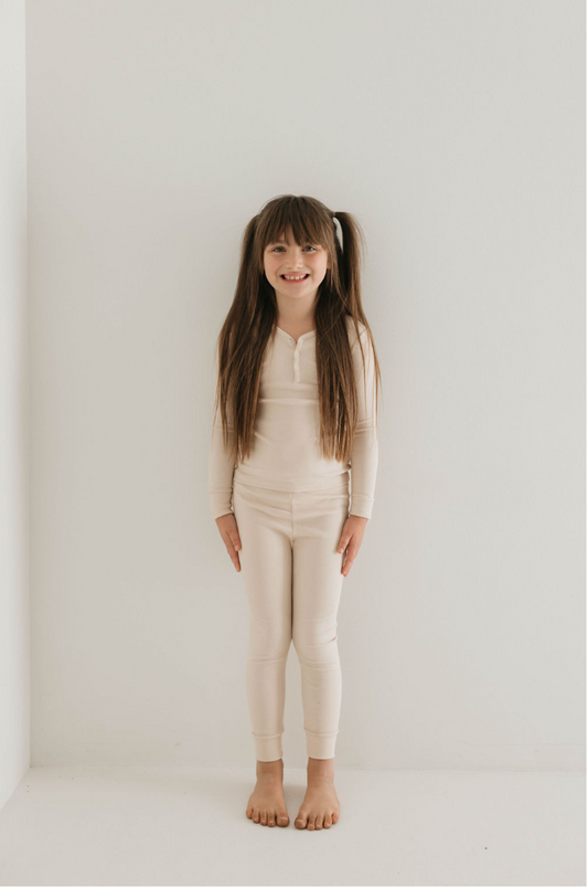 A young girl with long brown hair and bangs stands against a plain white background. She is wearing the Ribbed Cream Two Piece Bamboo Pajamas from forever french baby, which includes matching leggings. Barefoot and smiling brightly, her hair is tied in two playful ponytails.