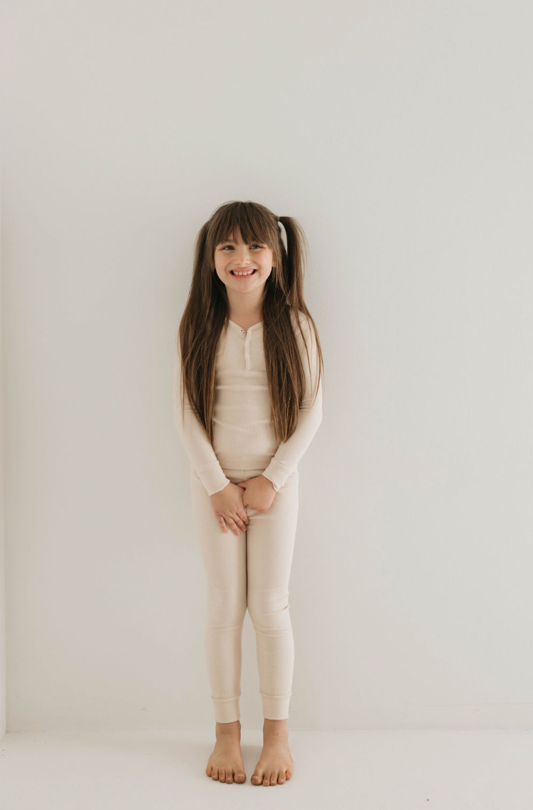 A young girl with long brown hair in pigtails stands smiling against a plain white background. She is wearing the "Two Piece Bamboo Pajamas | Ribbed Cream" by forever french baby, made from sustainable bamboo fabric and featuring matching leggings. Her ensemble looks as cozy as it is eco-friendly. With hands loosely clasped in front of her, she’s barefoot and adorable.