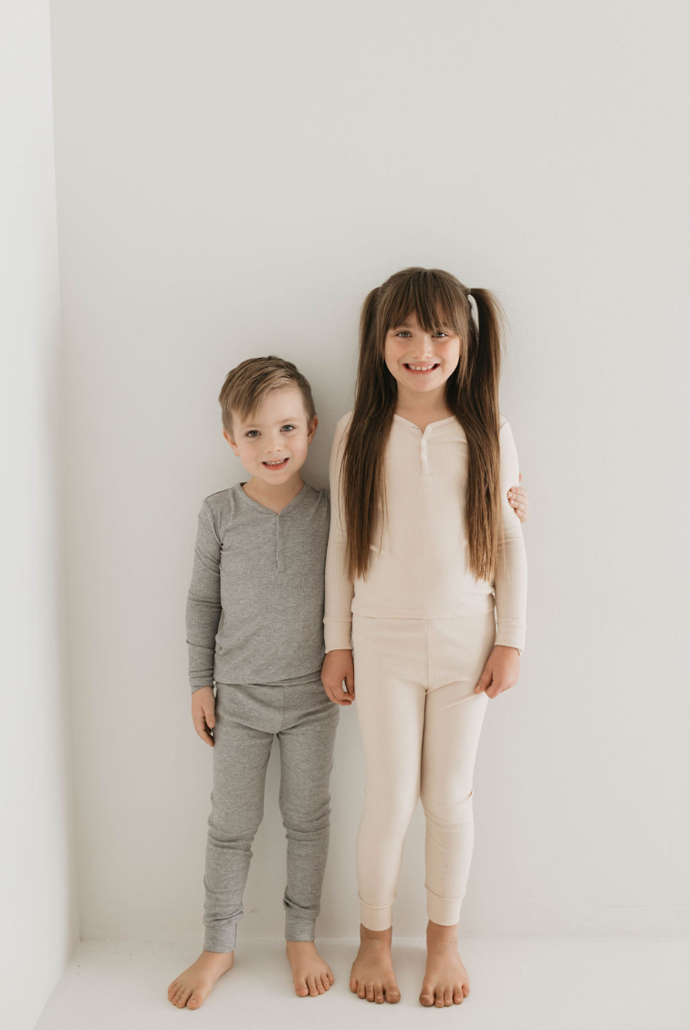 Against a plain white background, two children stand side by side. The child on the left is wearing grey bamboo pajamas, while the child on the right wears Two Piece Bamboo Pajamas | Ribbed Cream by forever french baby. Both are barefoot and smiling, with the child on the right's long hair tied into two ponytails.