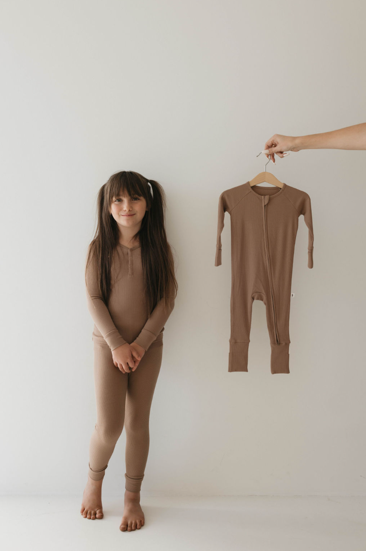 A young girl with long dark hair stands against a white wall wearing a ribbed milk chocolate outfit. To her right, an adult hand holds up smaller, identical Bamboo Zip Pajamas in Ribbed Milk Chocolate from forever french baby on a wooden hanger.