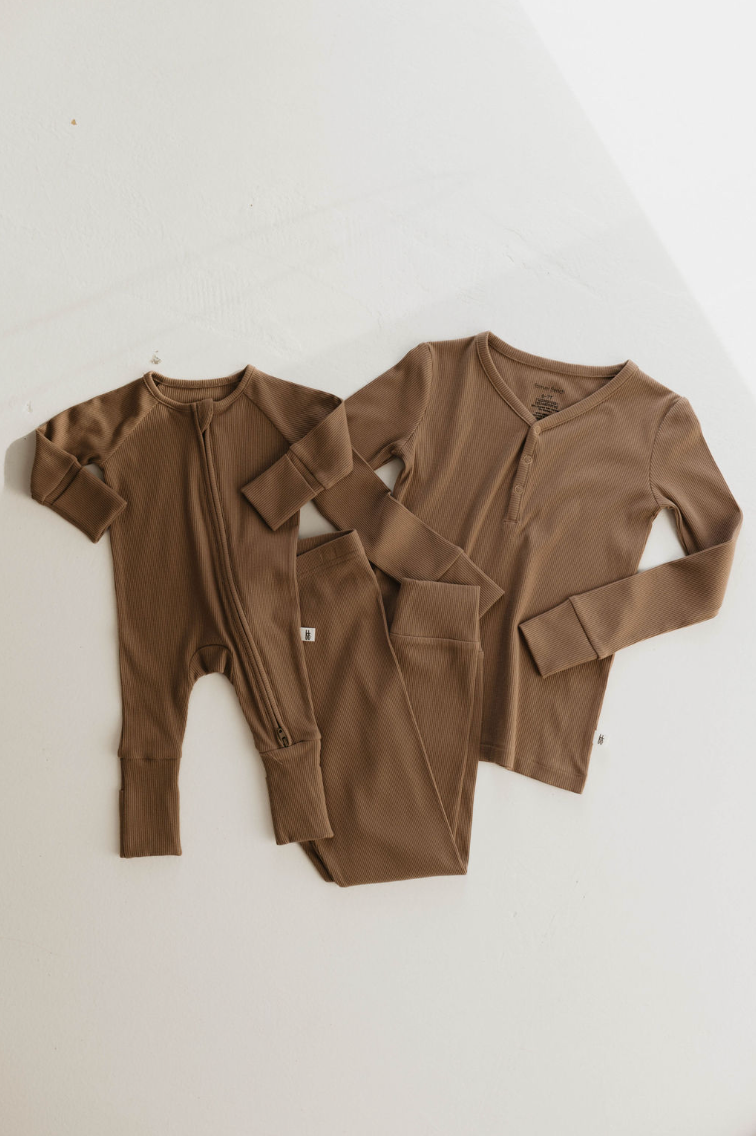 A baby onesie with a zipper and an adult long-sleeve top with pants, both in ribbed milk chocolate tones, are laid out on a white surface. Crafted from bamboo fabric by forever french baby, they exude a cozy, warm appearance.