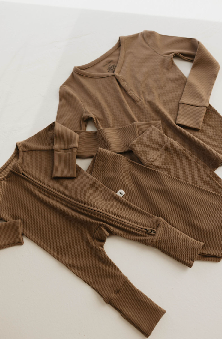 Two ribbed milk chocolate baby onesies from forever french baby are on a light surface. One has front button closure, the other a convenient bamboo zipper. Both offer long sleeves and cuffs for comfort and style.