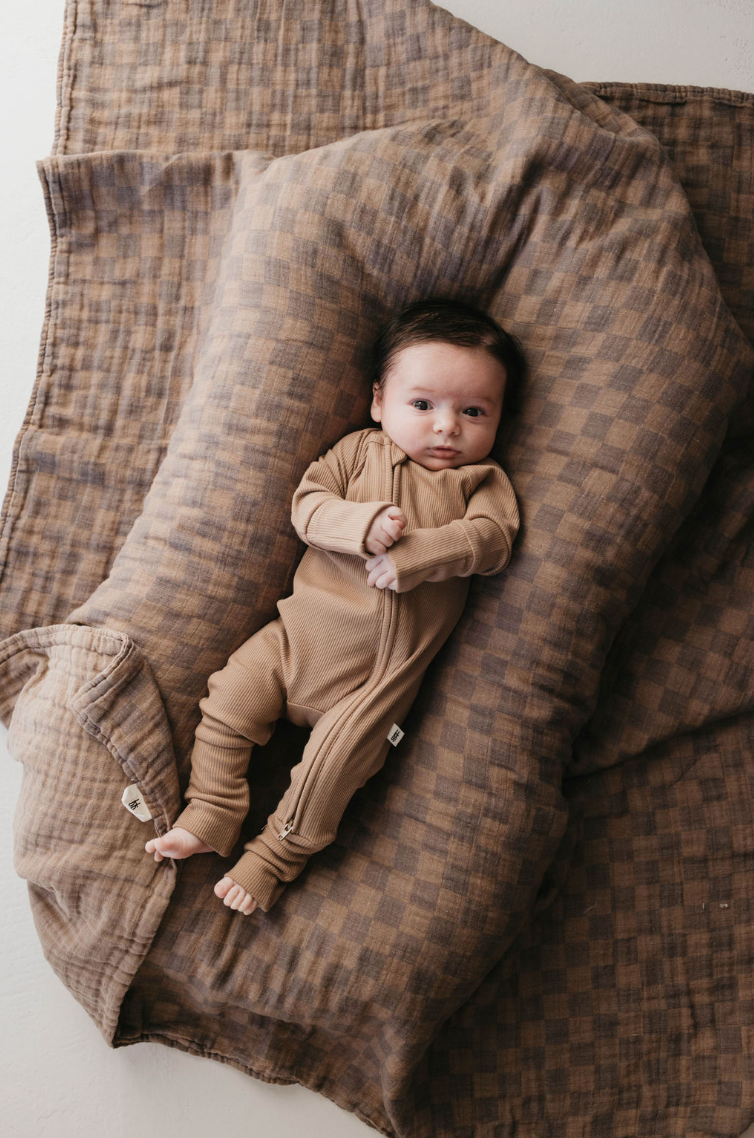 A baby in forever french baby's Bamboo Zip Pajamas, Ribbed Milk Chocolate, lies awake on a soft, checkered brown blanket. Tiny hands are visible near its face, making the cozy scene warm and inviting.