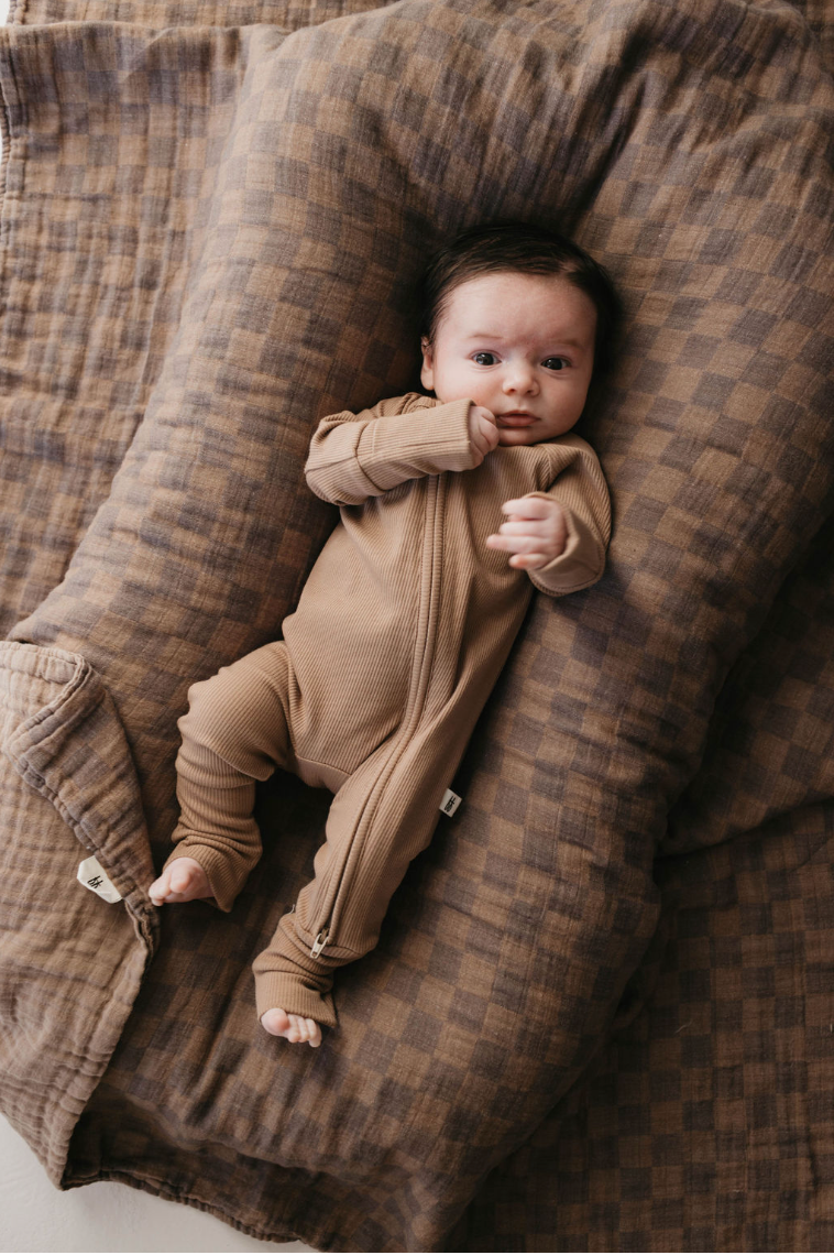 A baby in forever french baby's Bamboo Zip Pajamas, Ribbed Milk Chocolate, lies on a large, soft brown checkered blanket. With dark hair and a relaxed expression, the little one gently touches their cheek with one hand.