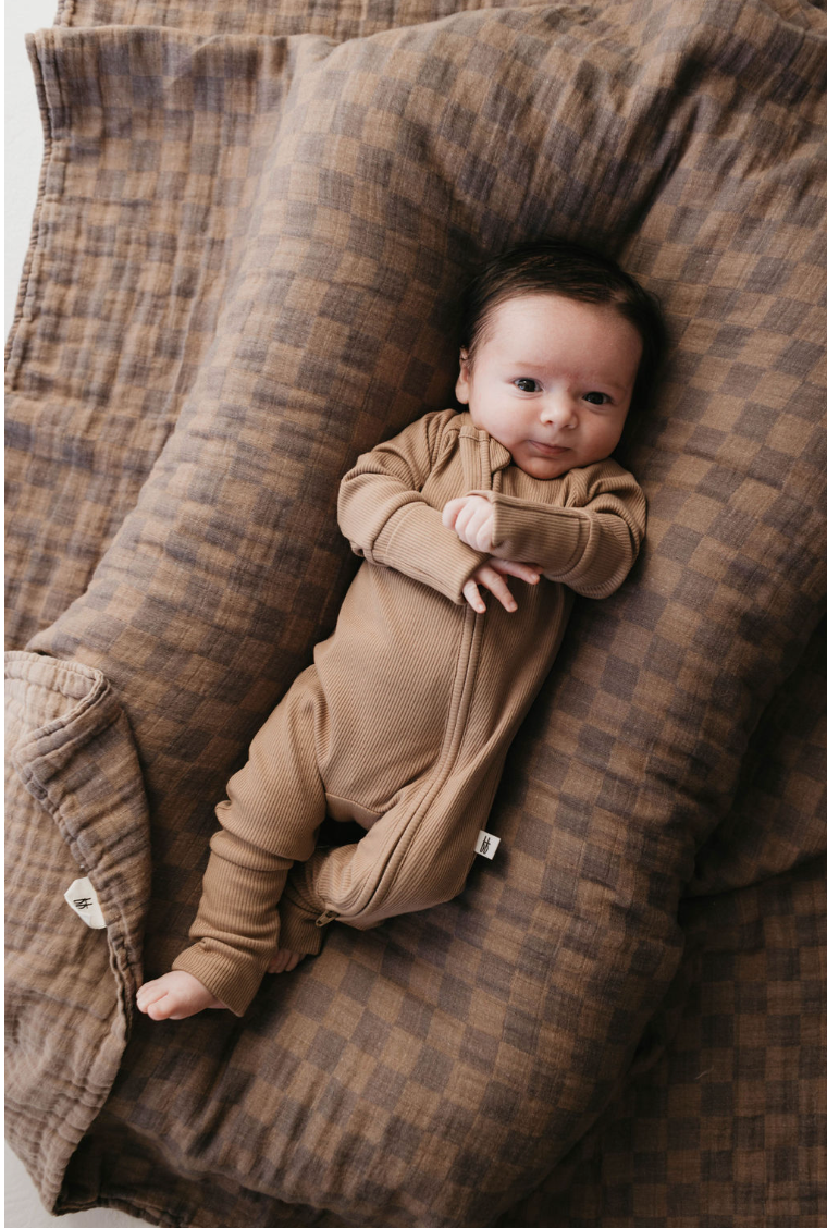 A baby in Bamboo Zip Pajamas (Ribbed Milk Chocolate) by forever french baby, with dark hair, rests on a plaid-patterned brown blanket in cozy, soft bedding, creating a warm and comforting scene.