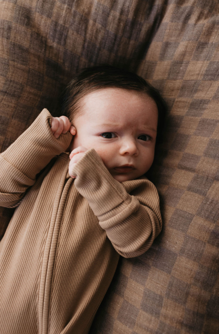 A baby in Forever French Baby's Bamboo Zip Pajamas in Ribbed Milk Chocolate lies on a brown plaid blanket, dark hair framing the curious face, and tiny hands nearby.