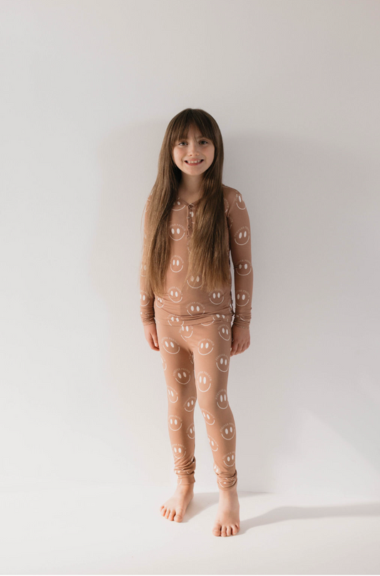 A child stands barefoot against a plain white background, wearing forever french baby's Bamboo Two Piece Pajamas in Desert Clay ff Smile, which feature a beige base color adorned with white smiley face patterns. The sustainable bamboo fabric promises a cozy night's sleep. They have long, straight hair and smile softly at the camera.