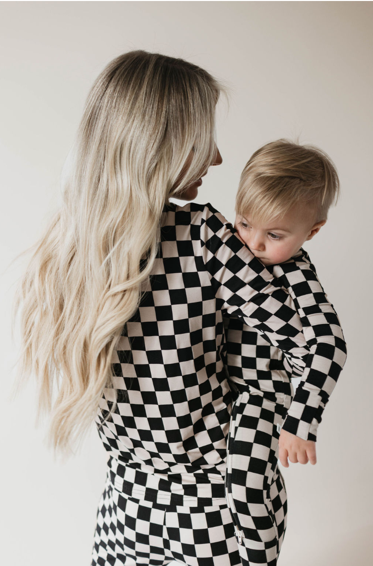 A woman with long, wavy blonde hair holds a young toddler. Both are dressed in forever french baby’s Women's Bamboo Pajamas in Black Checkerboard made of soft bamboo fabric. They are captured from the side against a plain background. The toddler looks to the side, leaning on the woman's shoulder.