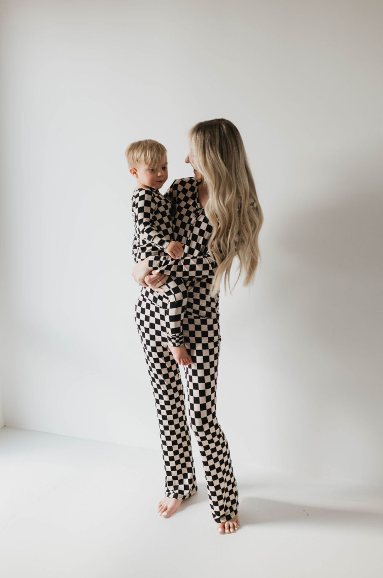 A woman with long blonde hair embraces a young child lovingly. They are both wearing matching Women's Bamboo Pajamas in Black Checkerboard by forever french baby, standing barefoot against a simple white background as they gaze at each other affectionately.