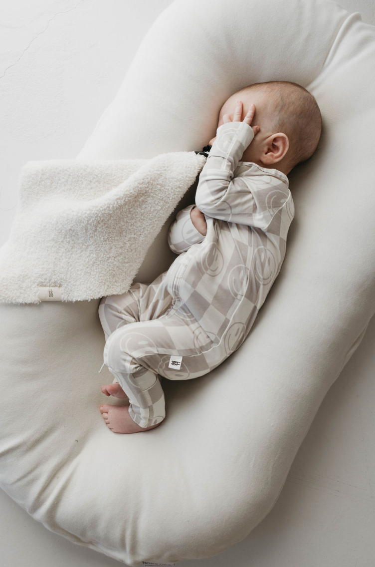 A baby wearing breathable Bamboo Zip Pajamas in the Smile Checkerboard pattern from forever french baby sleeps on a cushioned, sausage-shaped pillow. The baby has its hand covering its face and is partially covered with a white, fluffy blanket. The background is light-colored and minimalistic.