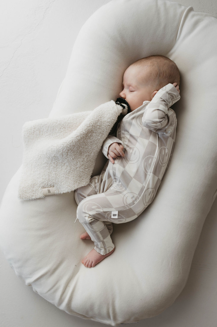 A baby sleeps on a cushioned white pillow, wearing hypo-allergenic Bamboo Zip Pajamas in the Smile Checkerboard pattern from forever french baby. The beige onesie adorned with smiley face patterns adds to the comfort, while the baby holds a small, white blanket close to their face, creating a cozy and serene sleeping environment.