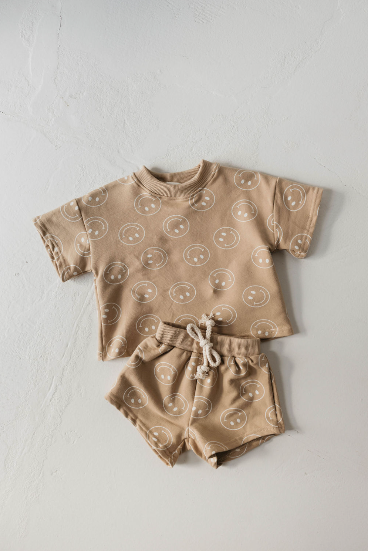 The "Short Set | Just Smile Tan" from forever french baby is laid flat against a light surface. This matching beige toddler set features a short-sleeved shirt and shorts, both adorned with a repeated pattern of simple, white smiley faces. The shorts include a white drawstring at the waistband, making it a charming addition to any children's wardrobe.