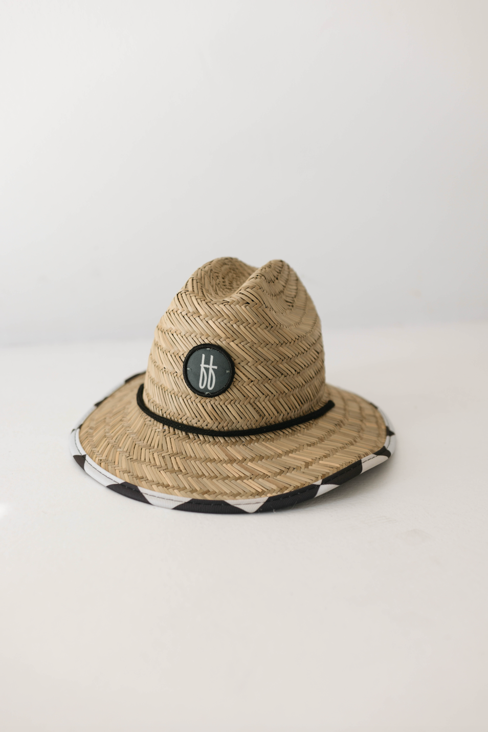 The "Straw Sun Hat | Black Checker" by forever french baby is a toddler sun hat featuring a black braided band around the base of the crown and a circular black patch showcasing the letters "bf" in white. The brim is accented with a distinct black-and-white patterned edge. This unisex beach hat, known for providing excellent sun protection, is displayed against a plain white background.