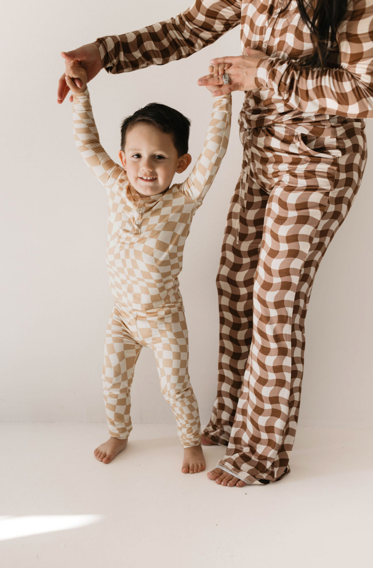 A young child wearing Groovy Gingham Bamboo Pajamas in beige and white checks beams with joy while being held up by an adult in matching brown and white checkered Women's Bamboo Pajamas from forever french baby. The adult's face is not visible in the photo, but they are standing in a brightly lit room with a light background that highlights the cozy bamboo fabric.