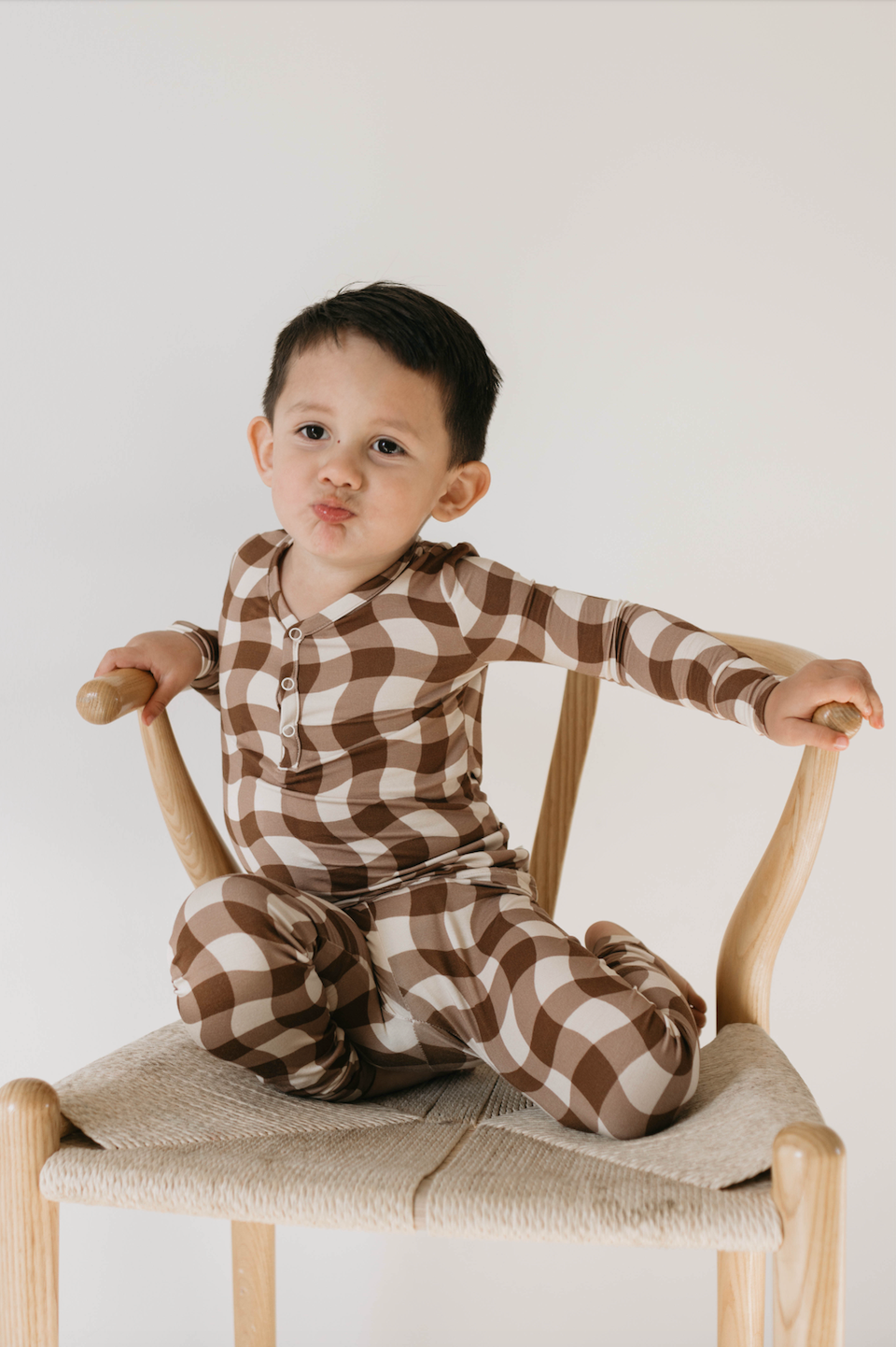 A young child with short dark hair is sitting on a wooden chair, wearing Bamboo Two Piece Pajamas in Groovy Gingham by forever french baby. The brown and white checkered outfit complements their playful puckering expression. The background is plain white.