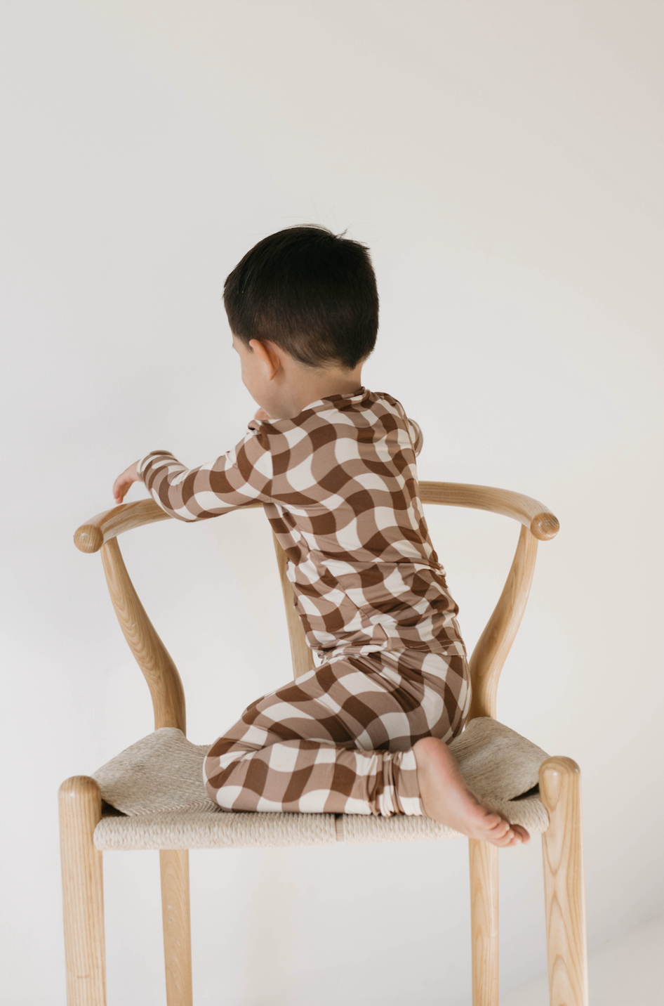 A young child with short dark hair, dressed in forever french baby's Bamboo Two Piece Pajamas in Groovy Gingham, sits backward on a wooden chair, facing away from the camera. The child is barefoot, with one foot resting on the chair seat and the other hanging off the edge. The background is plain white.