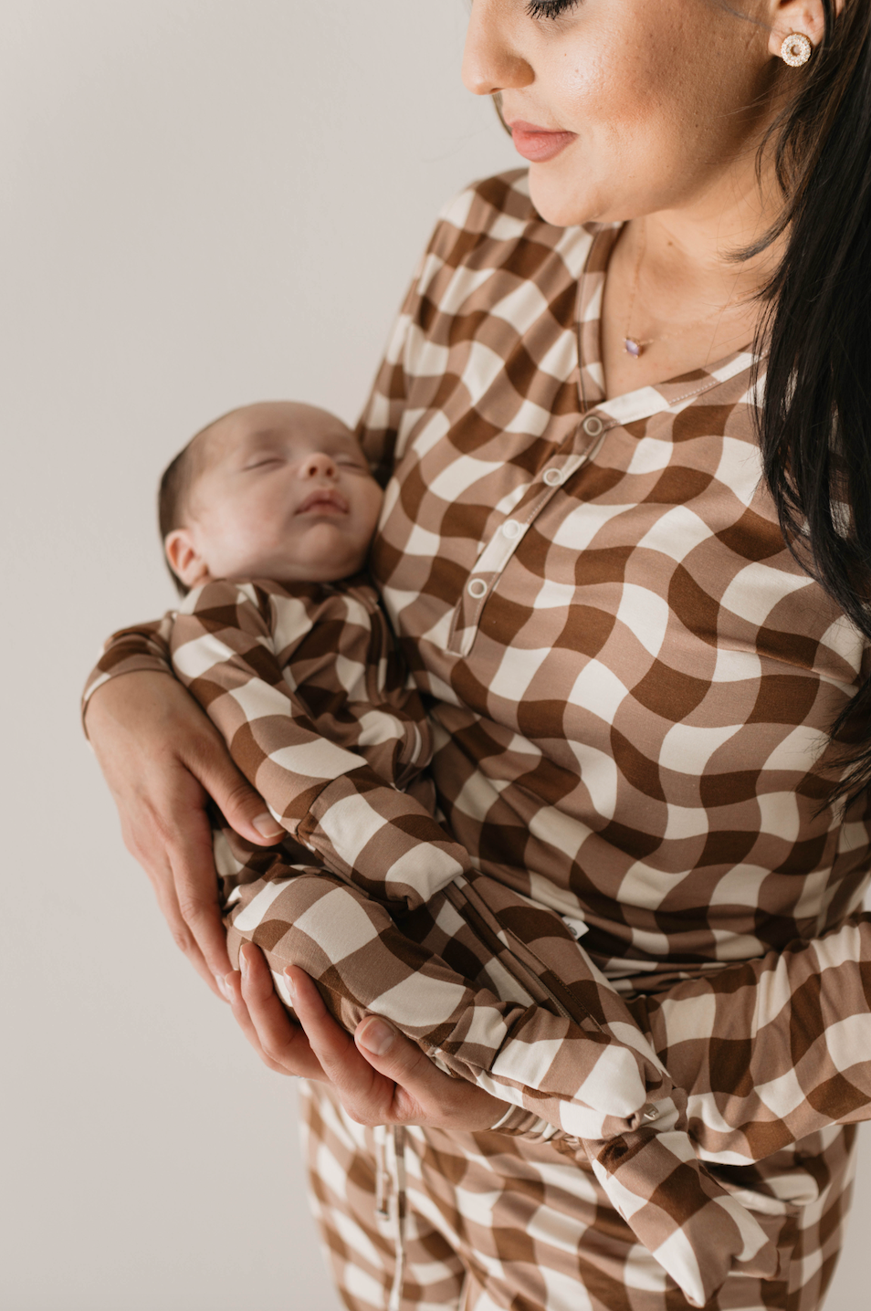 A woman holds a sleeping baby while both wear matching Groovy Gingham Women's Bamboo Pajamas from forever french baby. The woman, with long dark hair, gazes down at the baby with a serene expression. Made from bamboo fabric, these pajamas are ideal for breastfeeding moms. The background is plain white.