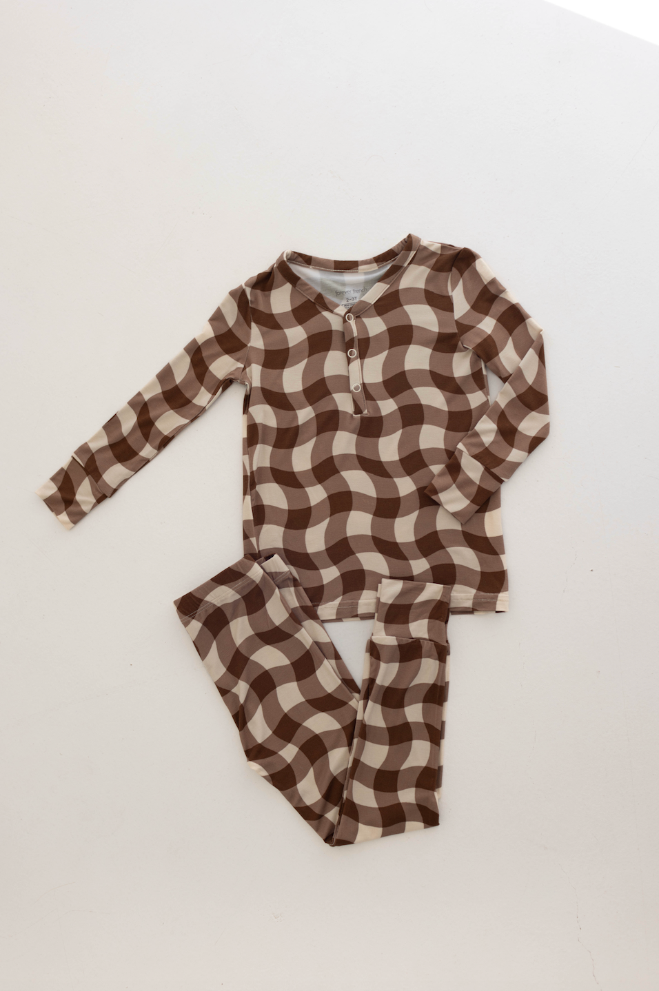 A set of Bamboo Two Piece Pajamas | Groovy Gingham from forever french baby, featuring a long-sleeved top and matching pants with a brown and beige checkered pattern. The top includes three small buttons near the neckline. These eco-friendly toddler pajamas are laid flat on a white surface.