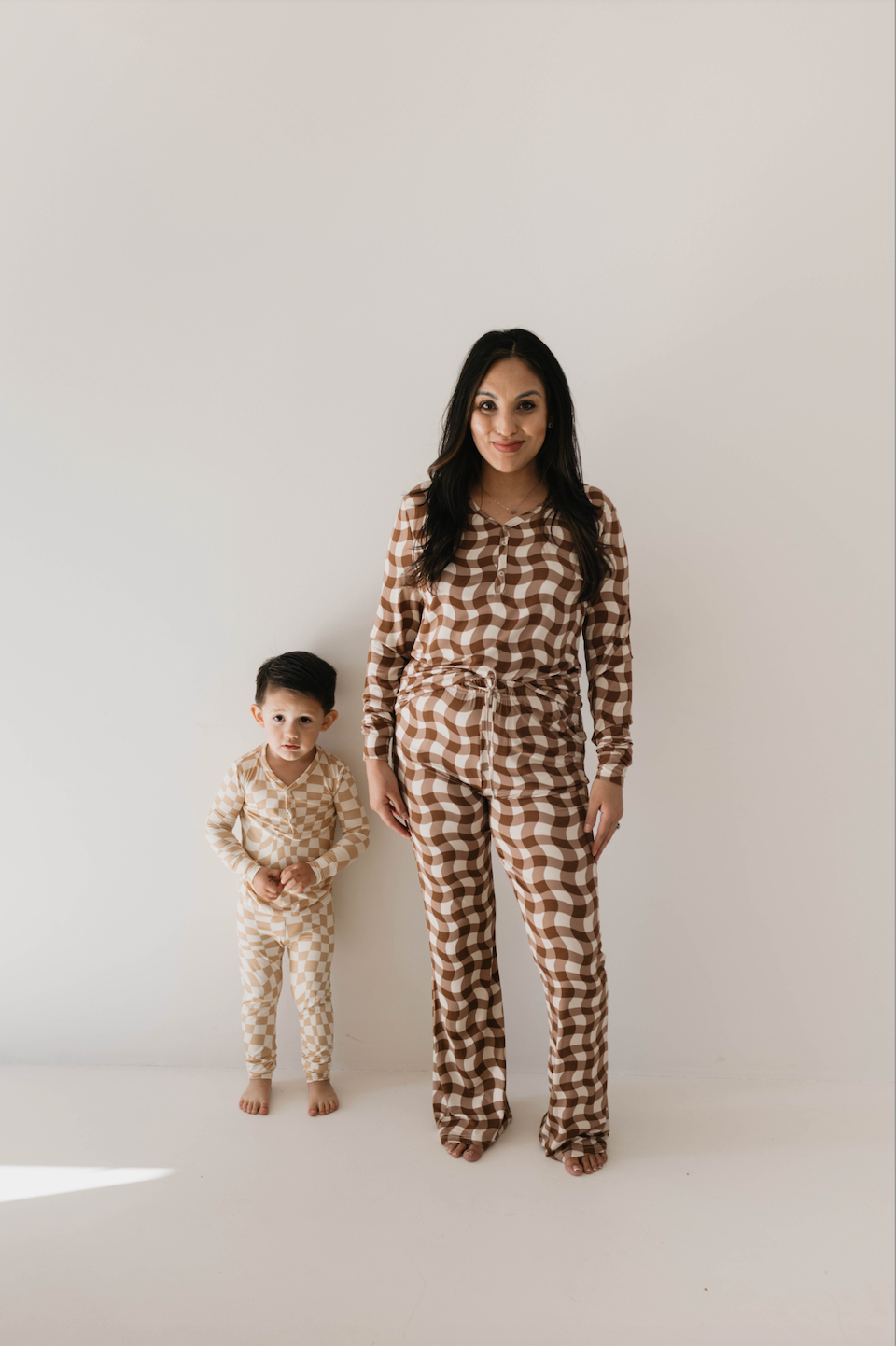 A woman and a young child stand side by side against a plain white background, both dressed in forever french baby's Groovy Gingham Bamboo Pajamas. The woman's outfit showcases brown and white checks, while the child's is adorned in lighter brown and white. They both look towards the camera, radiating coziness in their matching loungewear.