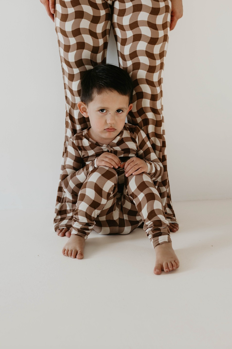 A young child dressed in forever french baby's Bamboo Two Piece Pajamas in Groovy Gingham sits on the floor looking somber, clad in matching brown and white checkered sleepwear. Leaning against an adult's legs also adorned with the same patterned pants, they create a poignant scene against a plain white background.