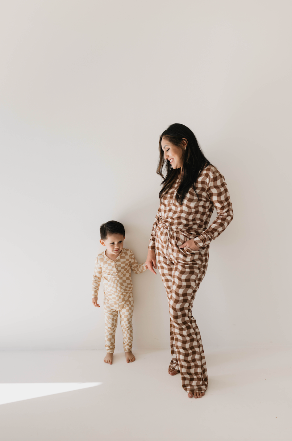 A woman and a young child stand against a plain white background. The woman is smiling and dressed in forever french baby's Women's Bamboo Pajamas | Groovy Gingham, while the child wears a similar outfit in a beige and white checkered pattern. They are holding hands and looking at each other, perfectly showcasing this comfortable bamboo fabric choice for breastfeeding moms.