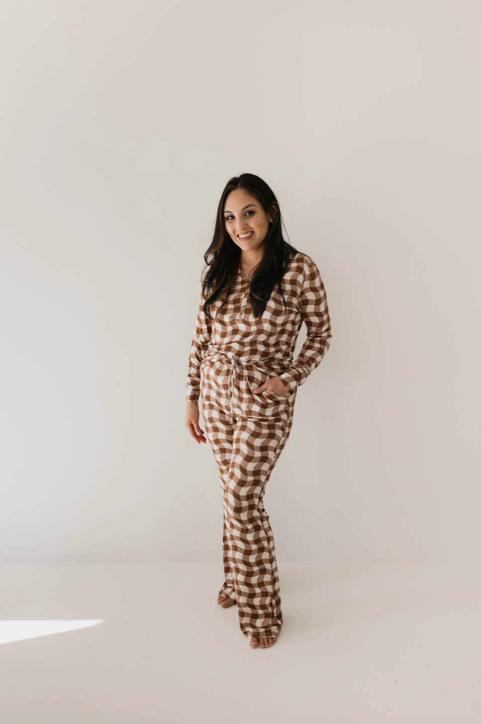 In front of a white backdrop, a woman wearing forever french baby's Women's Bamboo Pajamas in Groovy Gingham (brown and white) stands smiling with her right hand in her pocket. Her long dark hair flows freely as she strikes a relaxed pose, perfectly capturing the comfort of bamboo fabric ideal for breastfeeding moms.