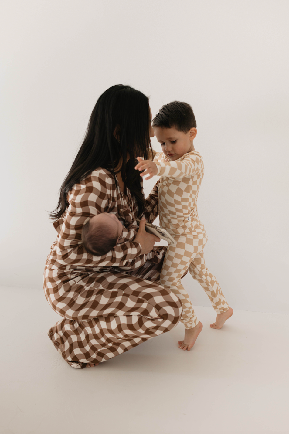 A woman with long dark hair is crouching down, holding a small baby wrapped in a brown checkered blanket. A young child in forever french baby's Women's Bamboo Pajamas | Groovy Gingham stands next to her with one arm outstretched, looking at the baby. They are in a minimalistic white space.